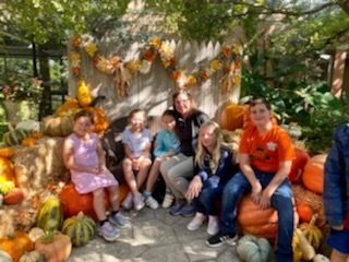third grade students with Mrs. Pletcher at sitting around pumpkins with a fence behind them and trees in the background